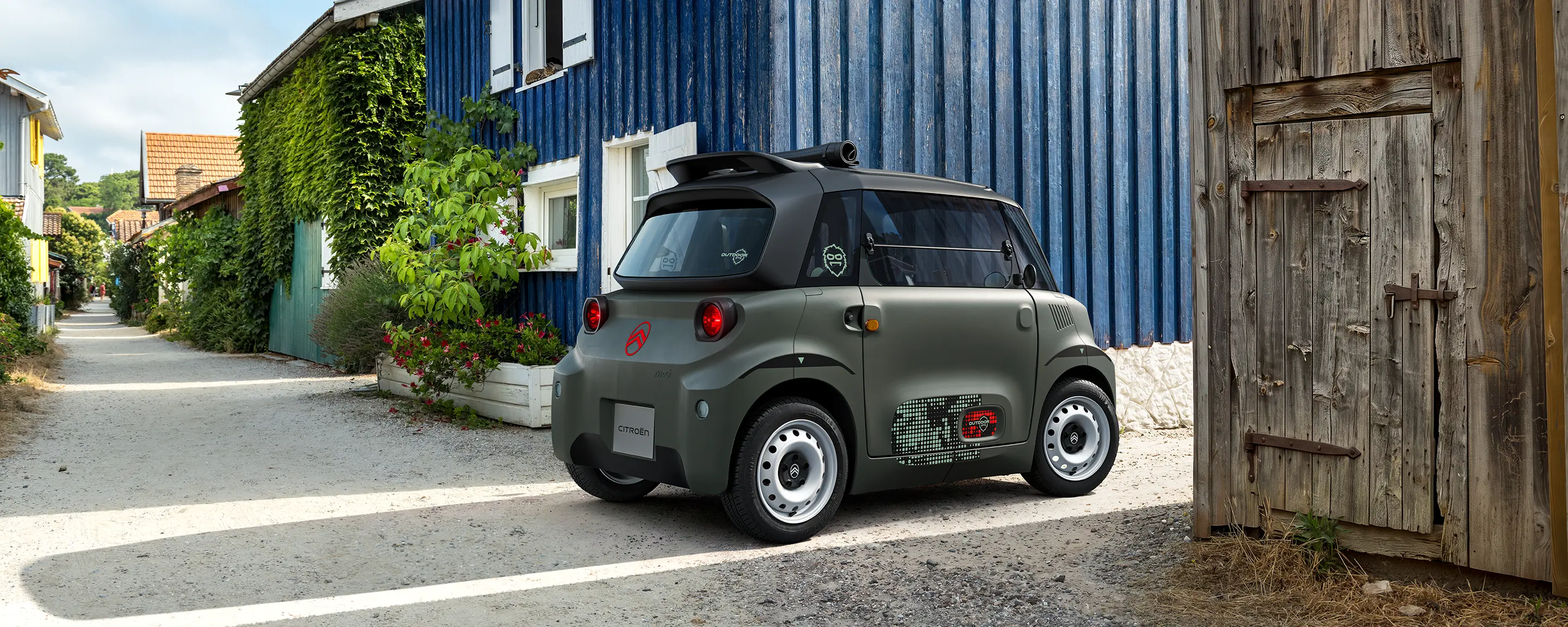 Vista de três quartos à frente: o Citroën ami Outdoor com faróis LED apresenta um automóvel elétrico urbano compacto numa vila costeira