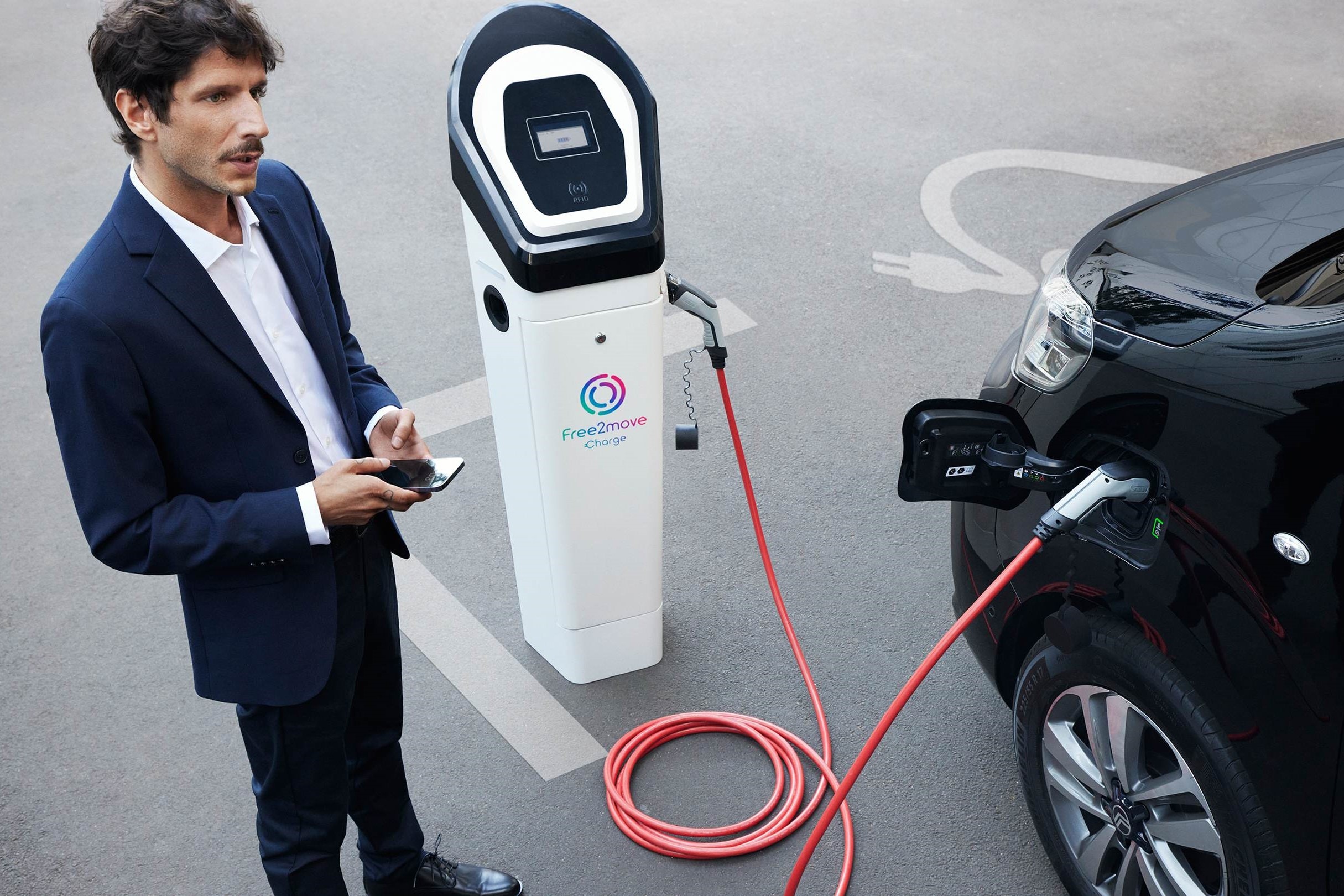 Veículo elétrico em carregamento numa estação pública enquanto uma aplicação no smartphone é utilizada para gerir o carregamento com o Free2move Charge